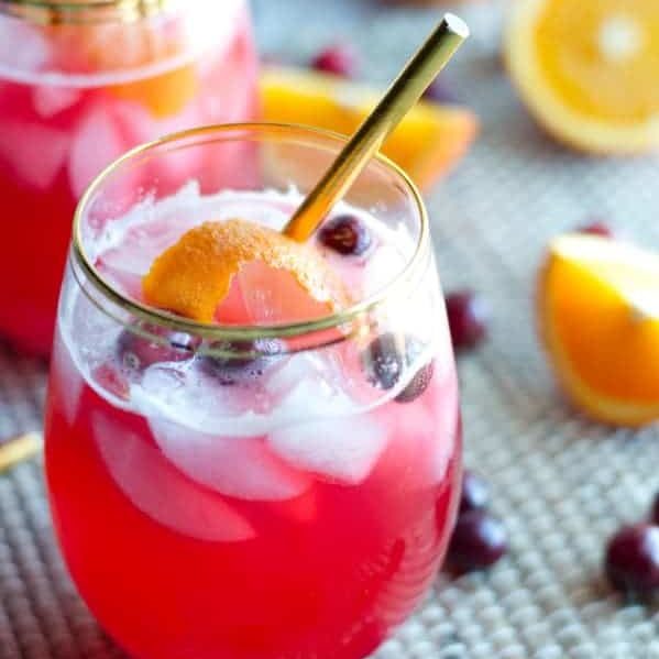 close up on glass and gold straw of vodka cranberry spritzer topped with ice, cranberries and orange peel