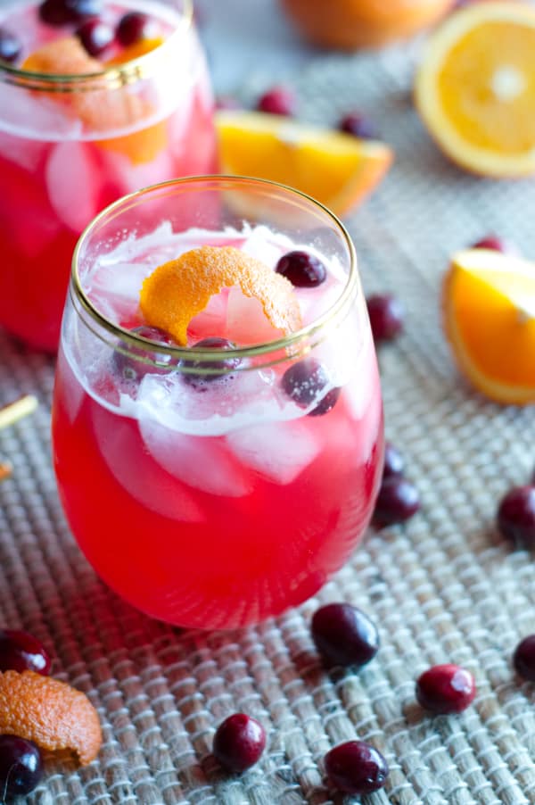 two glasses of vodka cranberry spritzer topped with ice, cranberries and orange peel. Whole cranberries and orange wedges scattered around on place mat