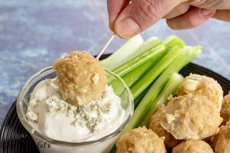 Hand holding toothpick skewered buffalo chicken meatball dipping into small bowl of blue cheese dip surrounded by platter of buffalo chicken meatballs and celery
