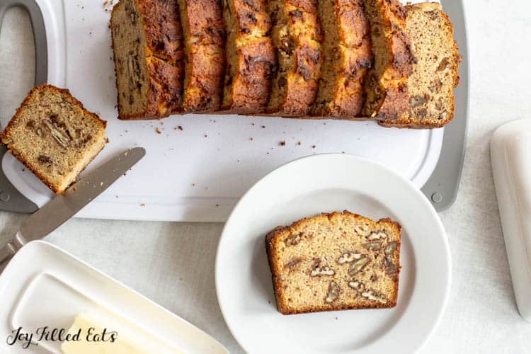 slices of keto banana bread on cutting board next to plate with single slice of banana bread. Butter knife and dish also next to cutting board and plate