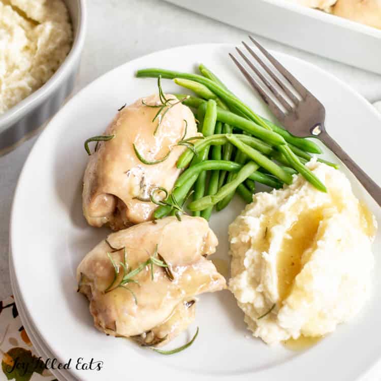 white plate of two baked rosemary chicken thighs served with a side of green beans and gravy covered mashed potatoes with fork