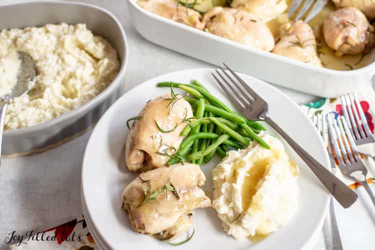 Baked Rosemary Chicken with a side of green beans and mashed potatoes on a white plate with a fork. Plate is next to a serving platter of mashed potatoes and casserole dish of baked rosemary chicken thighs