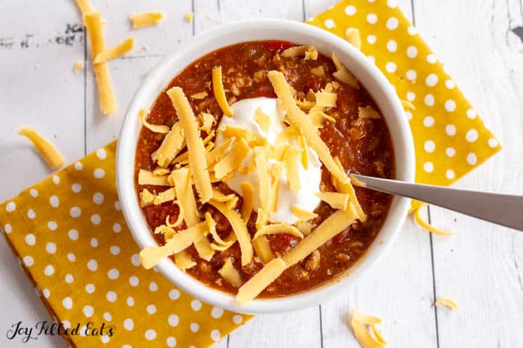 overhead view of instant pot turkey chili sprinkled with shredded cheese. Chili is in a white bowl with spoon on a yellow polka dot napkin.