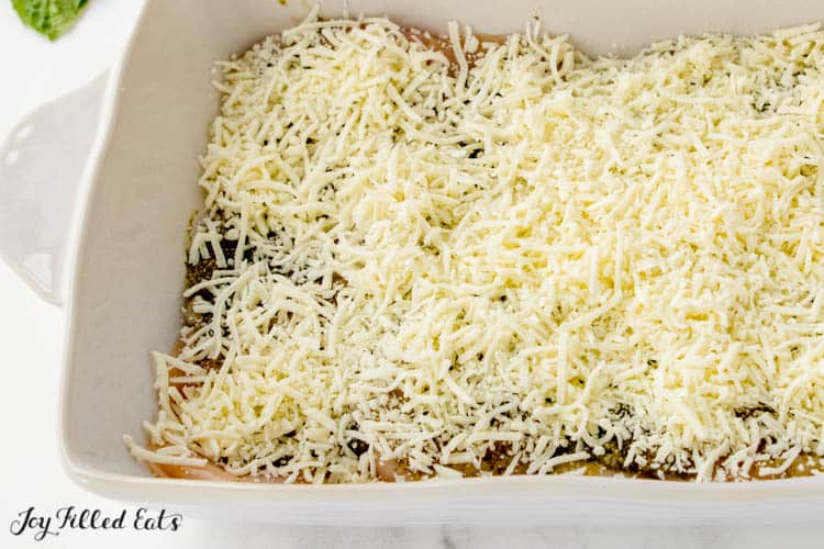 overhead view of white casserole dish with baked pesto chicken before baking, covered in shredded mozzarella cheese