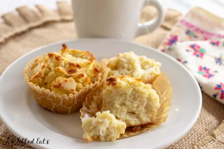 two coconut flour muffins in beige wrappers on white plate. One muffin is ripped in half. Plate is on beige place mat with decorative napkin and white mug