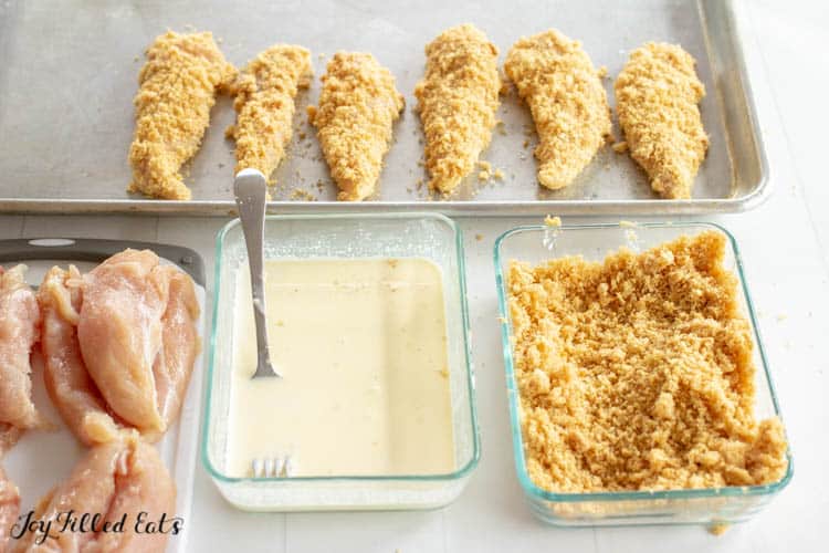 Sheet pan lined with precooked healthy baked chicken tenders next to raw chicken tenders, wet ingredients in a bowl with a fork and dry ingredient rub