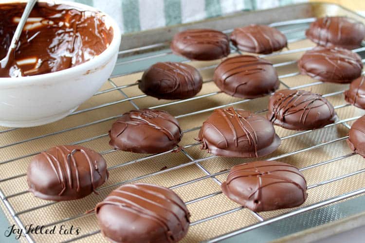 Homemade tagalong cookies on cooling rack covered in chocolate. Bowl of melted chocolate with spoon also on cooling rack