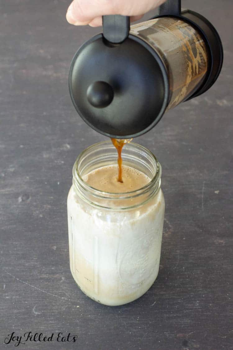 Hand pouring coffee from black french press into mason jar full of heavy cream and almond milk