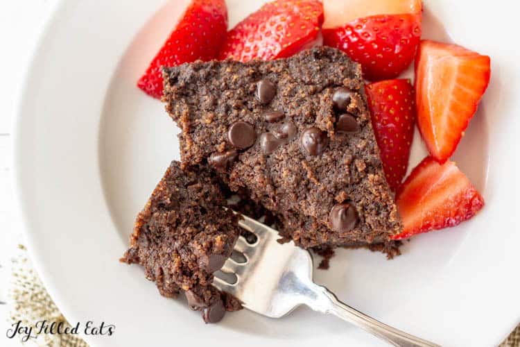 Overhead shot of brownie on a plate with small bite broken off on fork. Plate also includes a side of cut strawberries