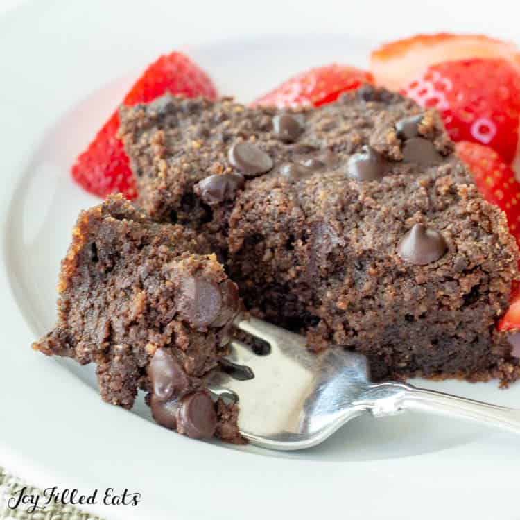 Close up of low carb brownie on a plate with small bite broken off on fork. Plate also includes a side of cut strawberries