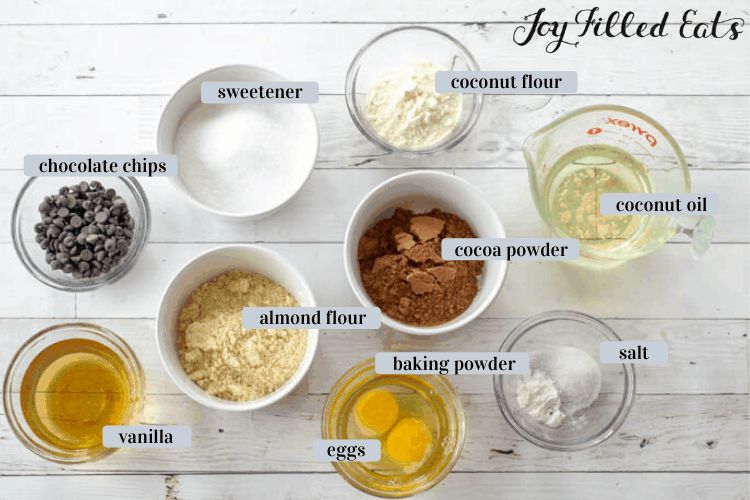 ingredients in various small bowls including chocolate chips, sweetener, coconut flour, coconut oil, cocoa powder, almond flour, vanilla, eggs, baking powder and salt