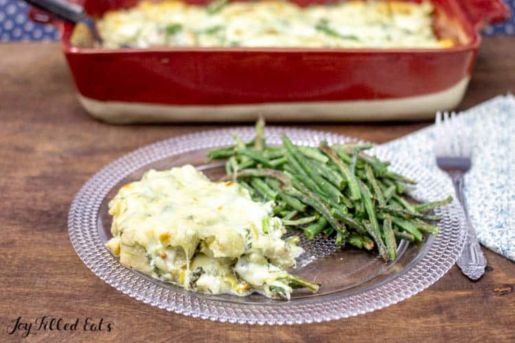 plate of spinach and artichoke chicken casserole served with a side of green beans. Fork on decorative napkin next to plate with red casserole dish in background