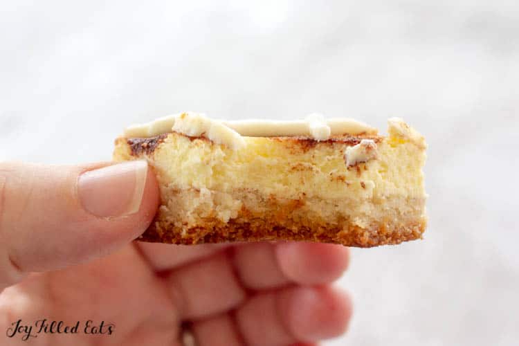 Hand holding cinnamon bun cheesecake bar in shape of square with bite taken out of piece