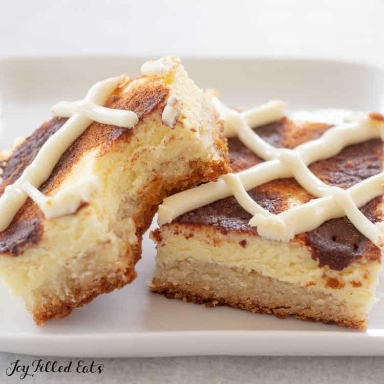 two cinnamon roll cheesecake bars on white plate with bite missing from one