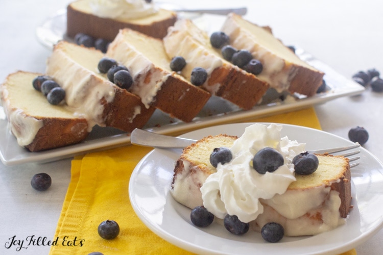 slice of lemon pound cake topped with whipped cream and blueberries on a plate in front of a platter of pound cake slices
