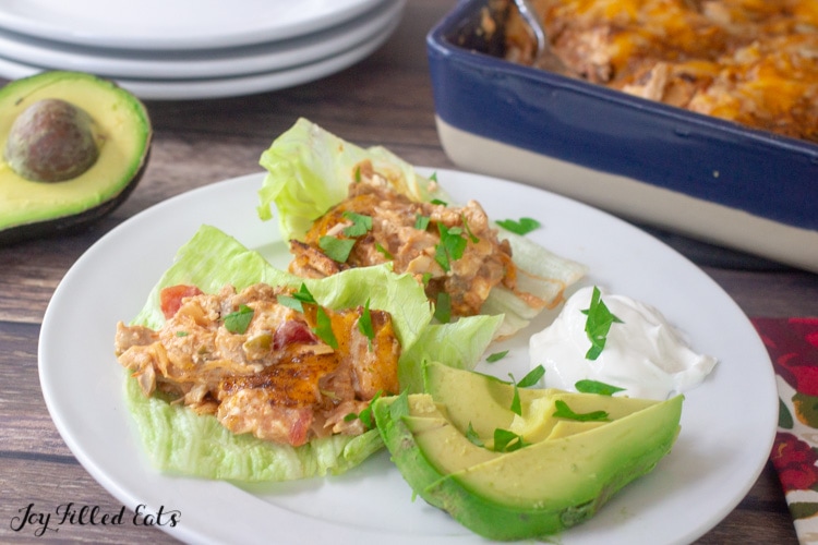 plate of Mexican chicken casserole servings on lettuce leafs next to sliced avocado and a dollop of sour cream. plate is set next to a halved avocado with the pit, stack of plates and casserole dish