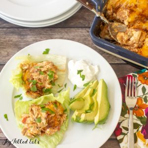 keto Mexican chicken casserole servings on lettuce leaves next to sliced avocado and a dollop of sour cream from above. plate is set next to a fork and decorative napkin, stack of plates and casserole dish with spoon.