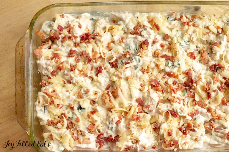 A Jalapeno Popper Chicken Casserole in a glass casserole dish with shredded cheese and bacon on top