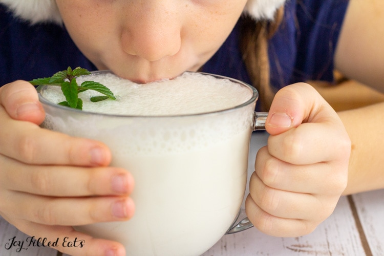 small child sipping from a glass of peppermint white hot chocolate topped with mint leaf