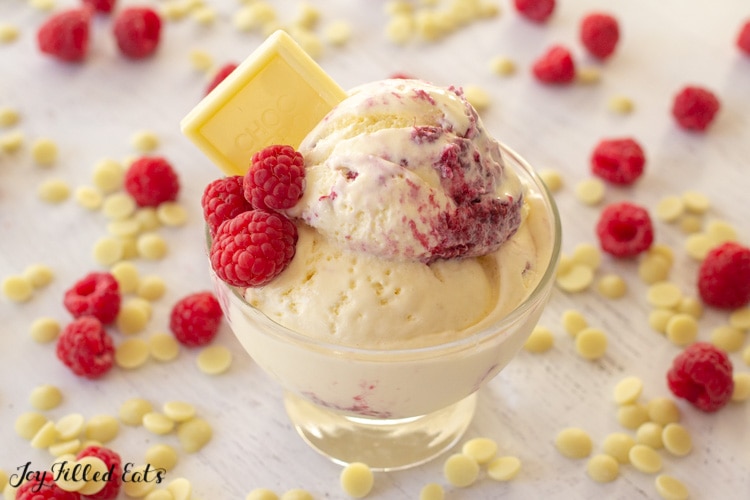 bowl of white chocolate raspberry ice cream topped with a white chocolate square and fresh raspberries with scattered white chocolate chips and berries around it