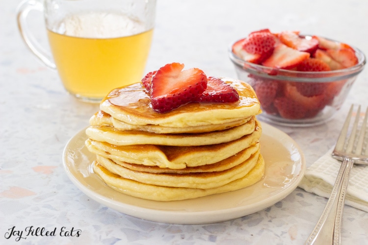 stack of pancakes in front of a pitcher of keto pancake syrup and a bowl of strawberries