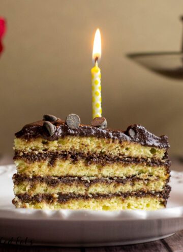 Slice of Keto Birthday cake layered with chocolate on a white plate. One lit yellow candle on the slice of cake.