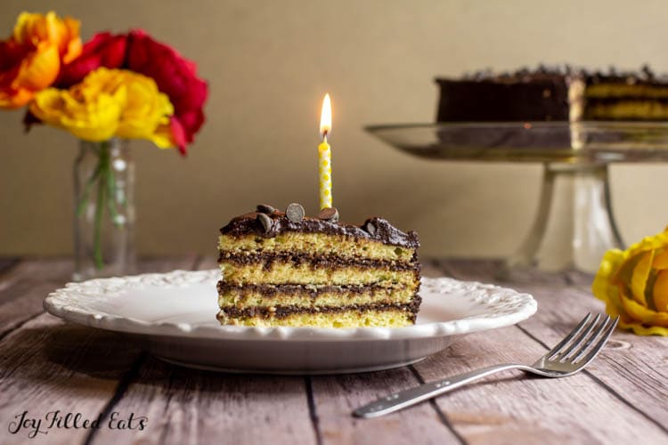 slice of yellow keto birthday cake with chocolate icing on a white plate with one lit yellow candle sticking in the slice. Plate is set next to a fork and cake platter with remaining cake