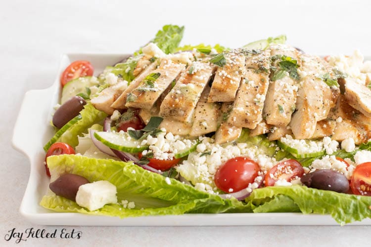 side view of green salad with grilled chicken, cucumbers, tomatoes, feta cheese, and kalamata tomatoes