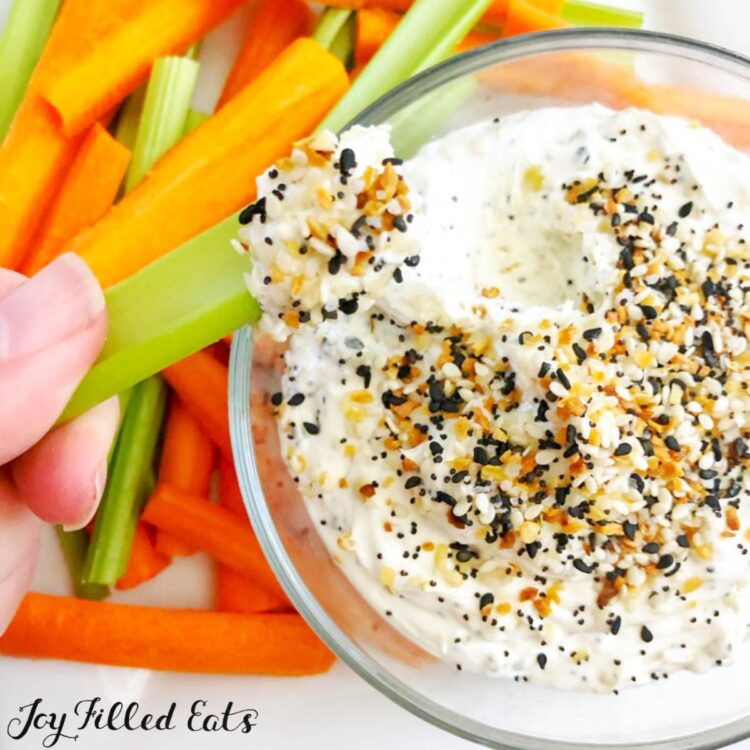 Hand holding celery stick with everything bagel dip on end in front of platter of celery and carrot sticks with a bowl of everything bagel dip
