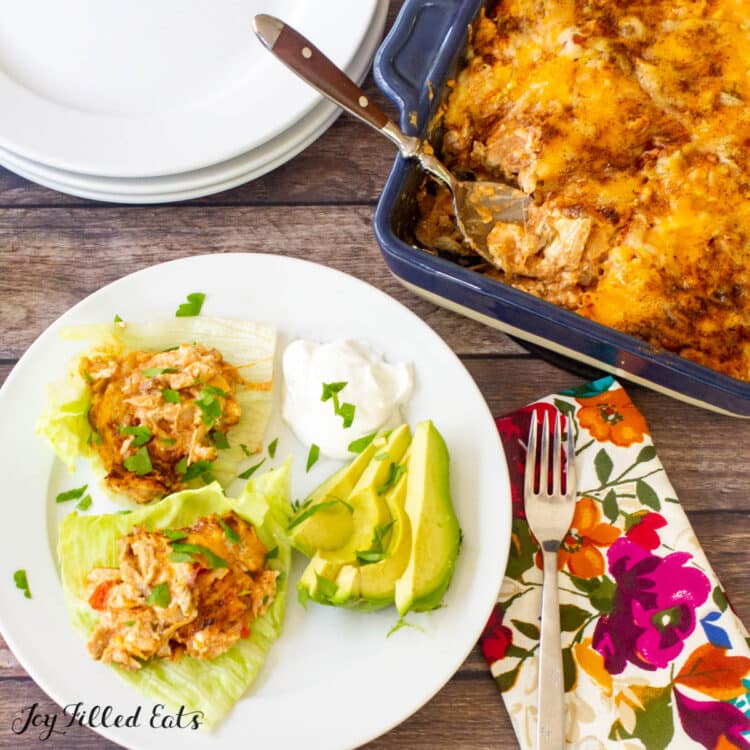 keto Mexican chicken casserole servings on lettuce leaves next to sliced avocado and a dollop of sour cream from above. plate is set next to a fork and decorative napkin, stack of plates and casserole dish with spoon.