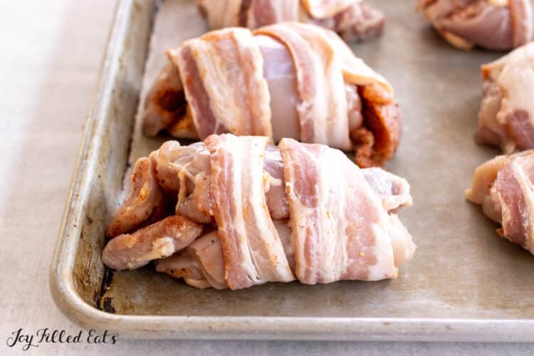 raw bacon wrapped around stuffed chicken thighs on baking pan