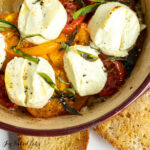baked goat cheese with tomatoes, basil, and onions shown close up in baking dish surrounded by sourdough toasts