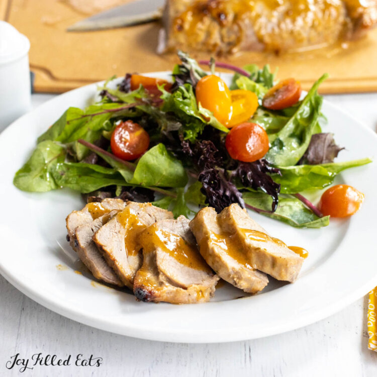 honey mustard pork tenderloin sliced on a dinner plate drizzled with honey mustard sauce with a side salad and remaining pork on cutting board behind