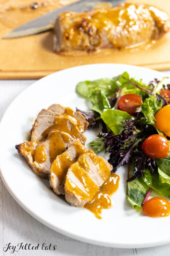 honey mustard pork tenderloin sliced on a dinner plate drizzled with honey mustard sauce with a side salad and remaining pork on cutting board behind