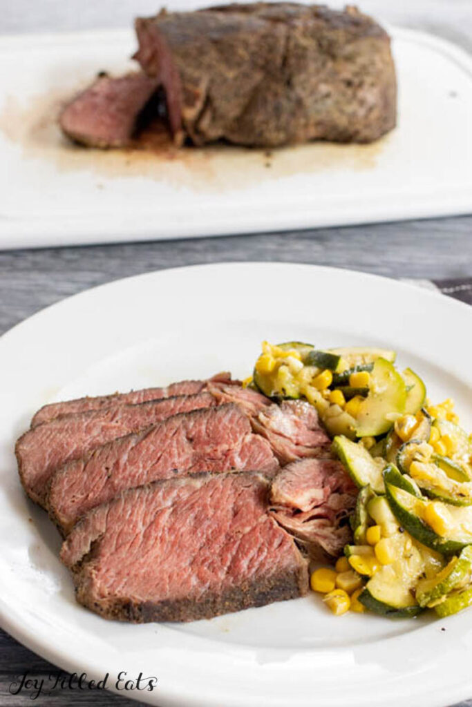 slices of sous vide beef chuck roast on dinner plate with veggies and cutting board behind